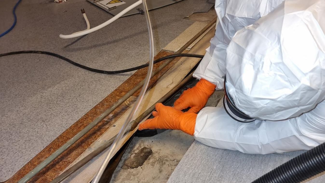 Person i vernedrakt arbeider med rør og kabler i gulv under rehabilitering innendørs